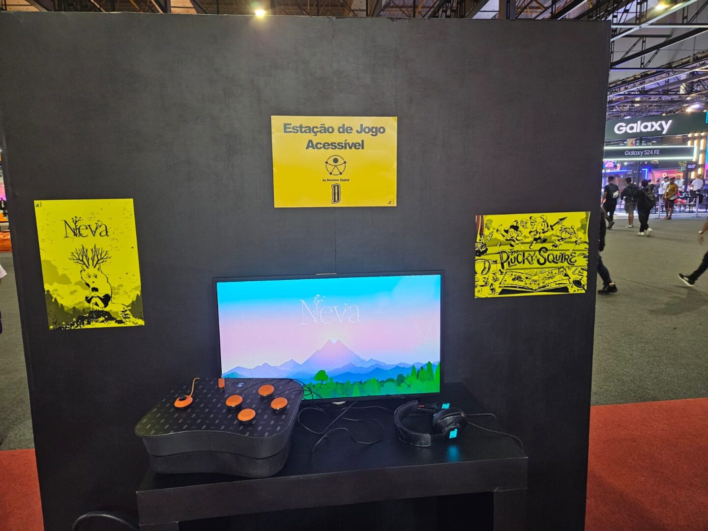 Imagem da Estação Acessível da AbleGamers no estande da Devolver Digital com o tela da TV com a tela de abertura do jogo Neva e em cima da bancada o controle YouMagic ao lado um headphone. na parede tem dois cartazes de Neva e The Plucky Squire ( oescudeiro Valente) em alto relevo e com alto contraste para que pessoas com baixa visão ou cegas possam interagir.